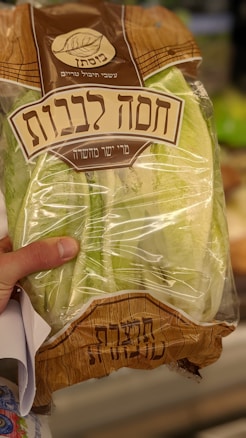 A bag of lettuce with Hebrew writing on the packaging is being held by a person's hand. The bag is transparent, allowing the fresh green leaves inside to be visible. The packaging has a brown design with a logo featuring a leaf.