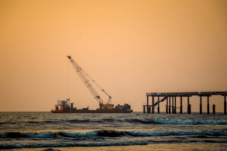 A tranquil seascape at sunset features a construction barge with a large crane positioned on the water. To the right, there is a partially constructed pier extending into the ocean. The sky is a warm orange hue, and gentle waves ripple across the water in the foreground.