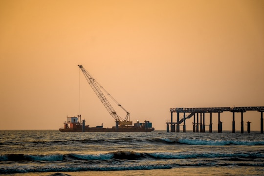 A tranquil seascape at sunset features a construction barge with a large crane positioned on the water. To the right, there is a partially constructed pier extending into the ocean. The sky is a warm orange hue, and gentle waves ripple across the water in the foreground.