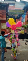 A street vendor is seen holding a bundle of colorful balloons and various toys in front of an entrance gate to a building. There is a yellow signboard above the entrance with text in an Indian language. The area is decorated with small green plants and signs, and several pairs of shoes are scattered on the ground near the entrance.