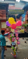 A street vendor is seen holding a bundle of colorful balloons and various toys in front of an entrance gate to a building. There is a yellow signboard above the entrance with text in an Indian language. The area is decorated with small green plants and signs, and several pairs of shoes are scattered on the ground near the entrance.
