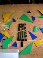 A brick wall decorated with colorful geometric shapes including triangles in blue, yellow, and green. In the center, stylized letters spell out 'PIC OLE Pops'. Below the letters is a small black vertical sign. A portion of a white surface, possibly a counter or freezer, is visible in the lower part of the image.