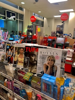 A store checkout area with magazines and various small products displayed near the counters. Prominent magazine covers feature well-known figures. Self-checkout stations are visible with 'Express Checkout' signs. The area is well-lit, and a few signs indicate available services like an ATM.