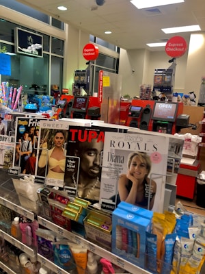 A store checkout area with magazines and various small products displayed near the counters. Prominent magazine covers feature well-known figures. Self-checkout stations are visible with 'Express Checkout' signs. The area is well-lit, and a few signs indicate available services like an ATM.