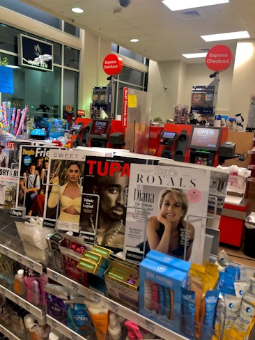 A store checkout area with magazines and various small products displayed near the counters. Prominent magazine covers feature well-known figures. Self-checkout stations are visible with 'Express Checkout' signs. The area is well-lit, and a few signs indicate available services like an ATM.