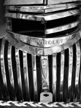 A vintage Chevrolet grill featuring horizontal chrome bars with signs of wear and a prominent emblem. The image has a classic and rugged appearance with visible dents and scratches.