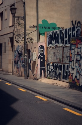 An urban scene with a narrow street lined by walls covered in vibrant graffiti. The artwork includes abstract designs, text in varying styles, and a stylized humanoid figure. Shadows cast by nearby buildings create contrasting patterns on the pavement.