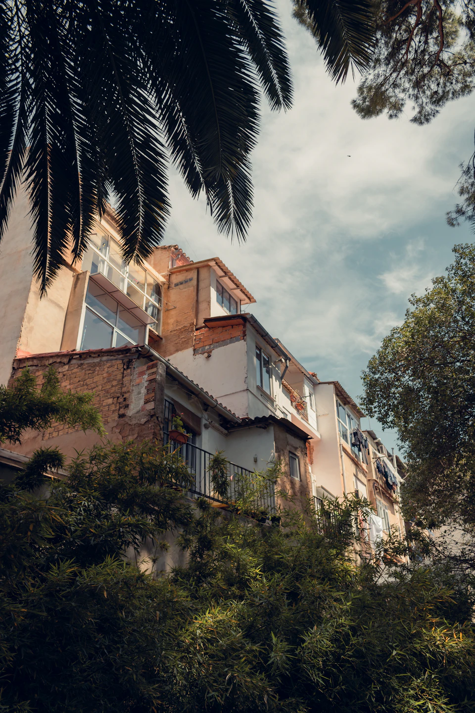 The image depicts a two-story building with white walls and red roofs, situated on a street corner surrounded by lush green trees. The perspective is from below, looking up at the building's facade, which adds to its imposing presence. The sky above is clear blue with scattered clouds, creating a serene backdrop for the urban structure.
