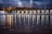 A vibrant lightning storm illuminates the night sky above a line of modern architectural structures along a riverside. The lightning bolts create dramatic reflections in the water, alongside reflections from streetlights. The scene is a juxtaposition of nature's raw power and urban development.