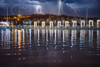 A vibrant lightning storm illuminates the night sky above a line of modern architectural structures along a riverside. The lightning bolts create dramatic reflections in the water, alongside reflections from streetlights. The scene is a juxtaposition of nature's raw power and urban development.