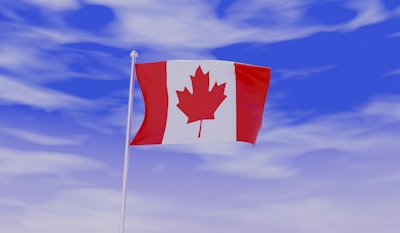 The Canadian flag with a red maple leaf at the center, flanked by two vertical red bands on a white background, is waving in a clear blue sky with scattered clouds.