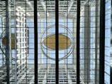A symmetrical architectural structure with a glass ceiling, featuring a large circular element in the center, surrounded by metal beams and glass panels. The background consists of a grid-like pattern with reflections and natural light, creating a sense of openness and modern design.