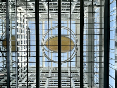 A symmetrical architectural structure with a glass ceiling, featuring a large circular element in the center, surrounded by metal beams and glass panels. The background consists of a grid-like pattern with reflections and natural light, creating a sense of openness and modern design.
