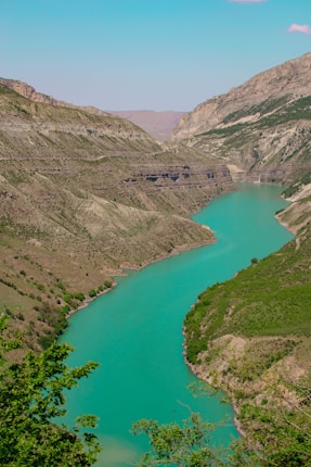 A winding river with vibrant turquoise water flows through a deep canyon surrounded by rocky hills. The lush greenery on the hill slopes contrasts with the arid mountaintops under a clear blue sky.