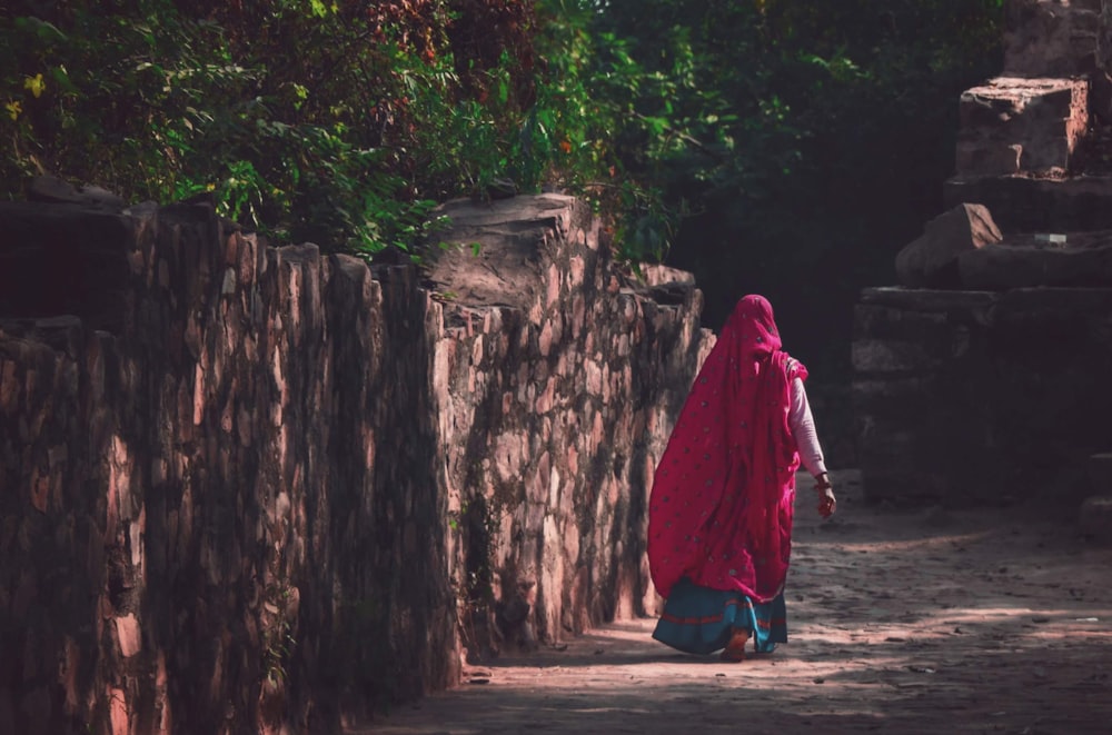 a woman walking down a dirt road next to a stone wall