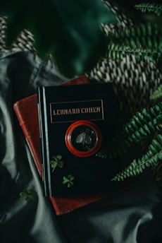 A vintage-style scene featuring a stack of books, with one prominently displaying the name 'Leonard Cohen'. The surroundings are accented with green leaves, including a fern and small clover-like plants. The overall composition includes dark and rich colors, creating a mysterious and moody atmosphere, with a magnifying glass resting on the top book.