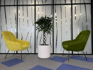 A modern office space featuring two upholstered chairs, one yellow and one green, positioned on opposite sides of a potted plant with a twisted trunk in a white planter. The background consists of frosted glass paneling with abstract vertical patterns, and the floor is carpeted with blue and gray tiles.