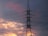 A tall electricity pylon stands silhouetted against a dramatic sky filled with clouds tinged with pink and orange hues. The delicate yet powerful presence of the power lines extends across the image, creating a striking contrast to the soft colors of the sunset.