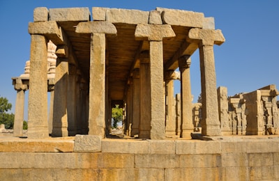 Ancient stone ruins with tall, intricately carved columns. The structure appears to be part of a historical site with detailed engravings on the pillars. Bright sunlight casts distinct shadows, enhancing the texture of the stone.
