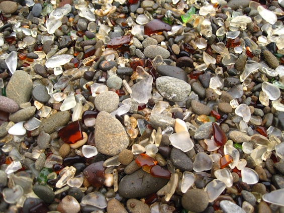 A collection of colorful pebbles mixed with small, smooth pieces of sea glass spread across a surface. The glass pieces are in various shades of brown, white, and green, and are scattered among the stones, which vary in size and color.