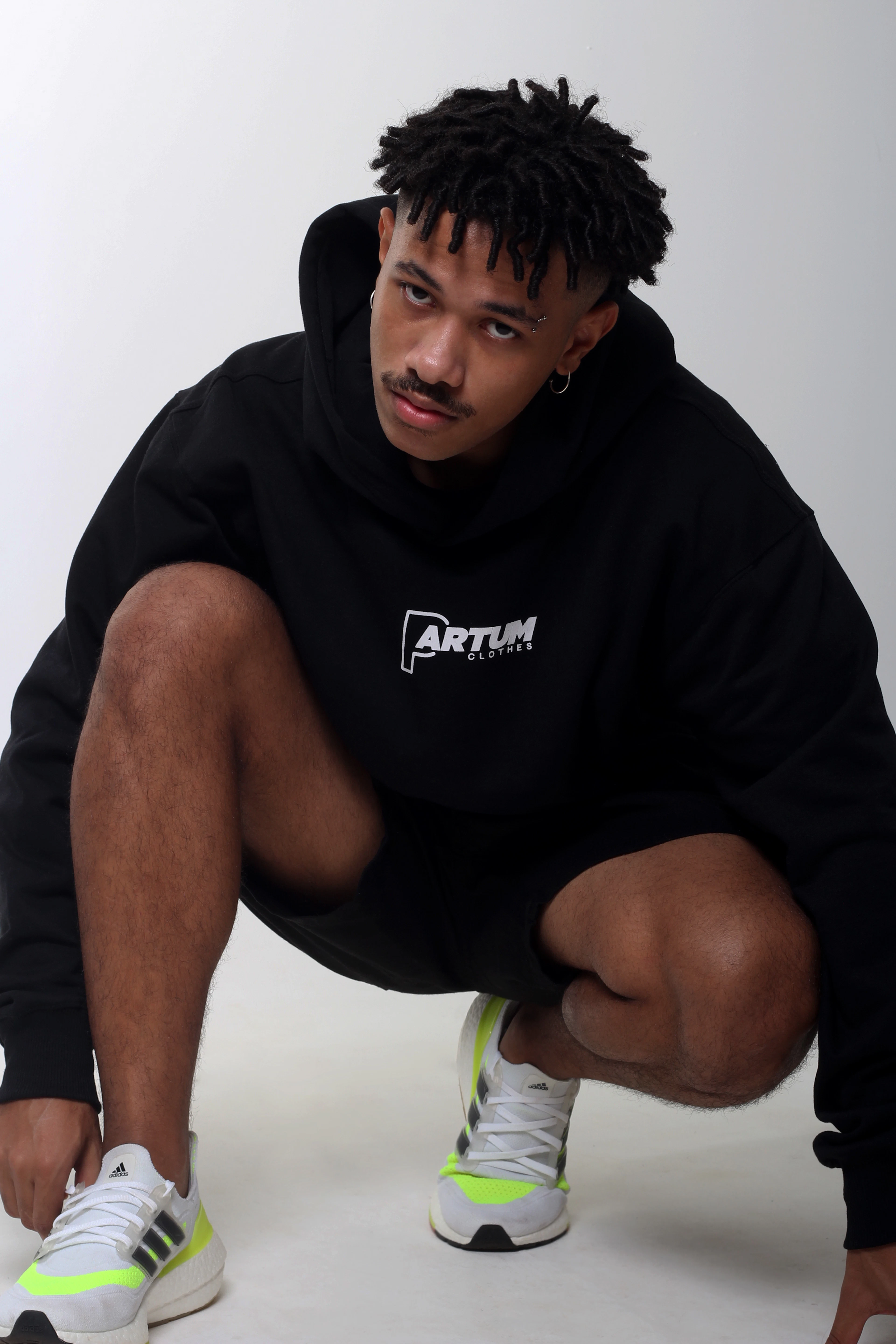 A man crouching in a black printed hoodie against a white studio wall.