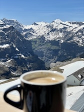 A black mug filled with coffee sits on a wooden table in the foreground. In the background, a stunning view of snow-capped mountains under a clear blue sky can be seen. The landscape features deep valleys and rocky peaks, with a small building partially covered in snow also visible to the side.