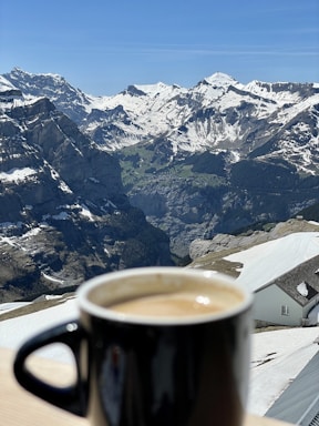 A black mug filled with coffee sits on a wooden table in the foreground. In the background, a stunning view of snow-capped mountains under a clear blue sky can be seen. The landscape features deep valleys and rocky peaks, with a small building partially covered in snow also visible to the side.