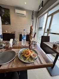 A dining table is set with a plate of fries garnished with sliced tomatoes and a side of sauce. The centerpiece is a vertical skewer with grilled meat. A bottle of water and glass cups are also on the table. The restaurant appears modern, with an air conditioner on the wall and outdoor light visible through the window.