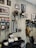 A room with a collection of vintage-style motorcycle helmets displayed on wall mounts and a shelf. There are framed black-and-white photographs and art prints on the walls, along with a lightbox sign saying 'Analog Klasik Est 2011'. Various accessories like wires and hooks hang on a grid organizer. A mirror is mounted on the wall above a mannequin head with a helmet. Jackets and a backpack hang from hooks, and there is a person sitting on a chair in the corner.