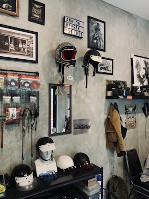 A room with a collection of vintage-style motorcycle helmets displayed on wall mounts and a shelf. There are framed black-and-white photographs and art prints on the walls, along with a lightbox sign saying 'Analog Klasik Est 2011'. Various accessories like wires and hooks hang on a grid organizer. A mirror is mounted on the wall above a mannequin head with a helmet. Jackets and a backpack hang from hooks, and there is a person sitting on a chair in the corner.