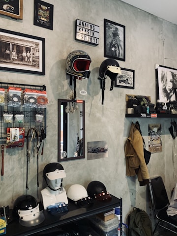 A room with a collection of vintage-style motorcycle helmets displayed on wall mounts and a shelf. There are framed black-and-white photographs and art prints on the walls, along with a lightbox sign saying 'Analog Klasik Est 2011'. Various accessories like wires and hooks hang on a grid organizer. A mirror is mounted on the wall above a mannequin head with a helmet. Jackets and a backpack hang from hooks, and there is a person sitting on a chair in the corner.
