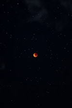 A dark night sky filled with stars surrounds a vivid red moon at the center, evoking a sense of mystery and tranquility. The moon's color suggests a lunar eclipse, creating a striking contrast against the darkness.