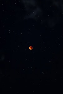 A dark night sky filled with stars surrounds a vivid red moon at the center, evoking a sense of mystery and tranquility. The moon's color suggests a lunar eclipse, creating a striking contrast against the darkness.
