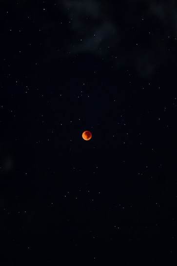 A dark night sky filled with stars surrounds a vivid red moon at the center, evoking a sense of mystery and tranquility. The moon's color suggests a lunar eclipse, creating a striking contrast against the darkness.