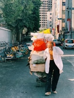 An elderly man is pulling a cart filled with various items, including bags and boxes, down a narrow urban street. The street is lined with bicycles and parked cars, while large buildings tower in the background. The scene is shaded by a large tree on the left.