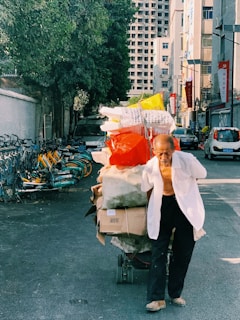 An elderly man is pulling a cart filled with various items, including bags and boxes, down a narrow urban street. The street is lined with bicycles and parked cars, while large buildings tower in the background. The scene is shaded by a large tree on the left.