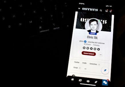 A smartphone screen displays a profile page with a black and white profile picture of a person. The screen indicates the status 'Founder' and shows social media icons and options like 'Remove Featured.' It is positioned in front of a dark keyboard, with illuminated keys slightly visible in the background.