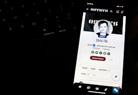 A smartphone screen displays a profile page with a black and white profile picture of a person. The screen indicates the status 'Founder' and shows social media icons and options like 'Remove Featured.' It is positioned in front of a dark keyboard, with illuminated keys slightly visible in the background.