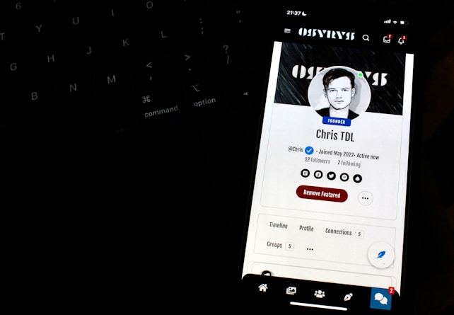 A smartphone screen displays a profile page with a black and white profile picture of a person. The screen indicates the status 'Founder' and shows social media icons and options like 'Remove Featured.' It is positioned in front of a dark keyboard, with illuminated keys slightly visible in the background.