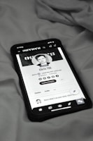 A smartphone displaying a social media profile screen rests on a fabric surface. The profile appears to belong to someone named Chris TDL with follow and following counts. The screen is centered in the image with a grayscale filter applied, and the edges of the phone case are visible.
