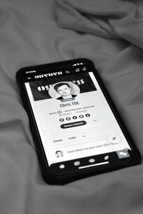 A smartphone displaying a social media profile screen rests on a fabric surface. The profile appears to belong to someone named Chris TDL with follow and following counts. The screen is centered in the image with a grayscale filter applied, and the edges of the phone case are visible.