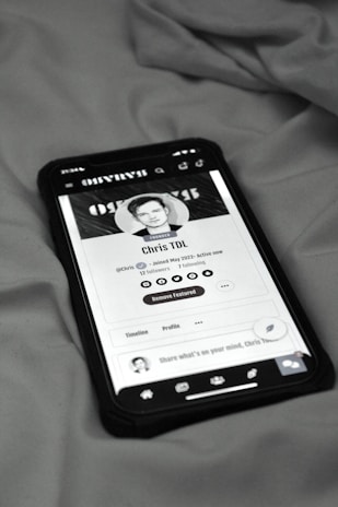 A smartphone displaying a social media profile screen rests on a fabric surface. The profile appears to belong to someone named Chris TDL with follow and following counts. The screen is centered in the image with a grayscale filter applied, and the edges of the phone case are visible.