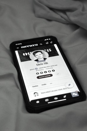 A smartphone displaying a social media profile screen rests on a fabric surface. The profile appears to belong to someone named Chris TDL with follow and following counts. The screen is centered in the image with a grayscale filter applied, and the edges of the phone case are visible.