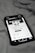 A smartphone displaying a social media profile screen rests on a fabric surface. The profile appears to belong to someone named Chris TDL with follow and following counts. The screen is centered in the image with a grayscale filter applied, and the edges of the phone case are visible.