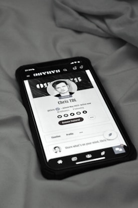 A smartphone displaying a social media profile screen rests on a fabric surface. The profile appears to belong to someone named Chris TDL with follow and following counts. The screen is centered in the image with a grayscale filter applied, and the edges of the phone case are visible.