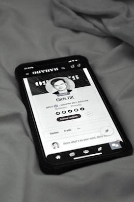 A smartphone displaying a social media profile screen rests on a fabric surface. The profile appears to belong to someone named Chris TDL with follow and following counts. The screen is centered in the image with a grayscale filter applied, and the edges of the phone case are visible.