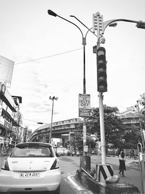 A busy urban street scene featuring a white car with a visible license plate stopped at a red traffic light. A railway bridge traverses the background, with a train traveling on it. Several pedestrians and cyclists are visible, and a street sign advertises a study program for IELTS and OET exams.