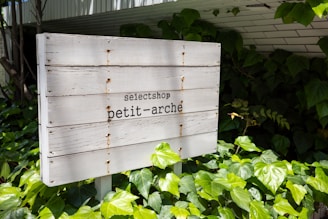 A wooden sign is standing amidst lush green leaf plants. The sign is slightly weathered and reads 'selectshop petit-arché' in black letters. The background includes more plants and a partially visible structure with a white tiled roof.