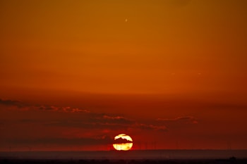 The image captures a vivid sunset with the sun partially visible on the horizon, casting a warm, orange glow across the sky. Dark clouds are scattered, adding contrast to the fiery sky. Wind turbines are faintly silhouetted against the horizon, creating a harmonious blend of nature and technology.