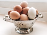 A collection of eggs sits in a metal colander on a light-colored surface. The colander has two handles and features small holes scattered across its surface. The eggs vary in color, with most being brown and one distinctly white.